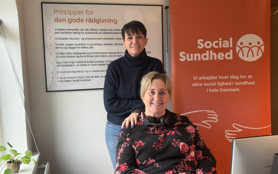 Ditte Ulv Hermansen bagerst med rådgiver Laila siddende i forgrunden. Bag ved dem hænger en plakat og orange roll-up